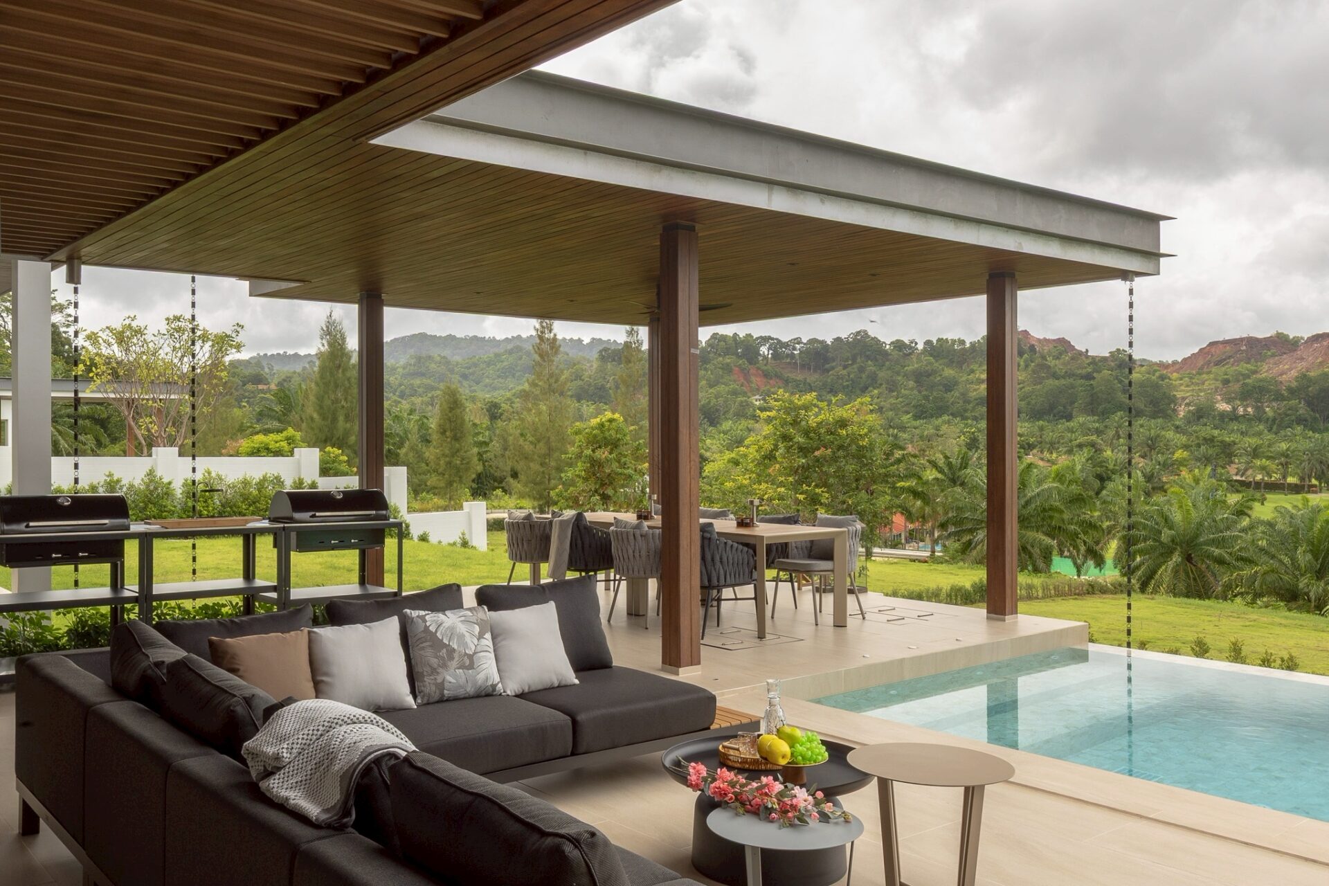 Outdoor covered patio with a dark sectional sofa, coffee tables, and a rectangular pool, overlooking lush green hills.