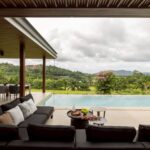 Outdoor patio with black cushioned seating beside a pool, table with drinks, and a lush, hilly landscape in the distance under cloudy skies.”