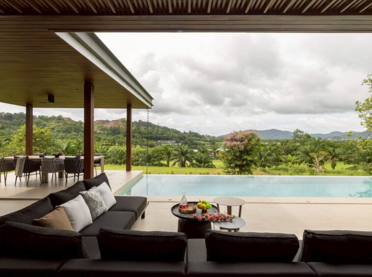 Outdoor patio with black cushioned seating beside a pool, table with drinks, and a lush, hilly landscape in the distance under cloudy skies.”