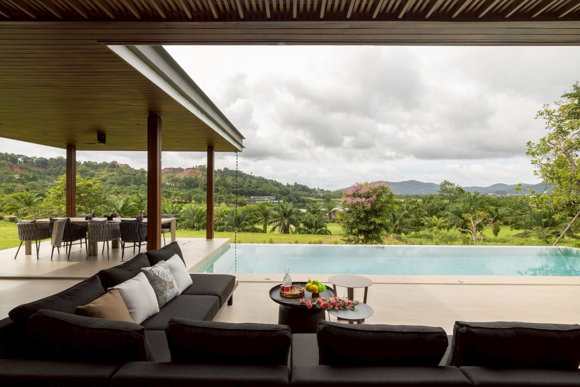Outdoor patio with black cushioned seating beside a pool, table with drinks, and a lush, hilly landscape in the distance under cloudy skies.”