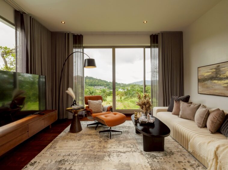 Modern living room with a beige sectional sofa, gray cushions, and large windows showing green hills outside on a sunny day.