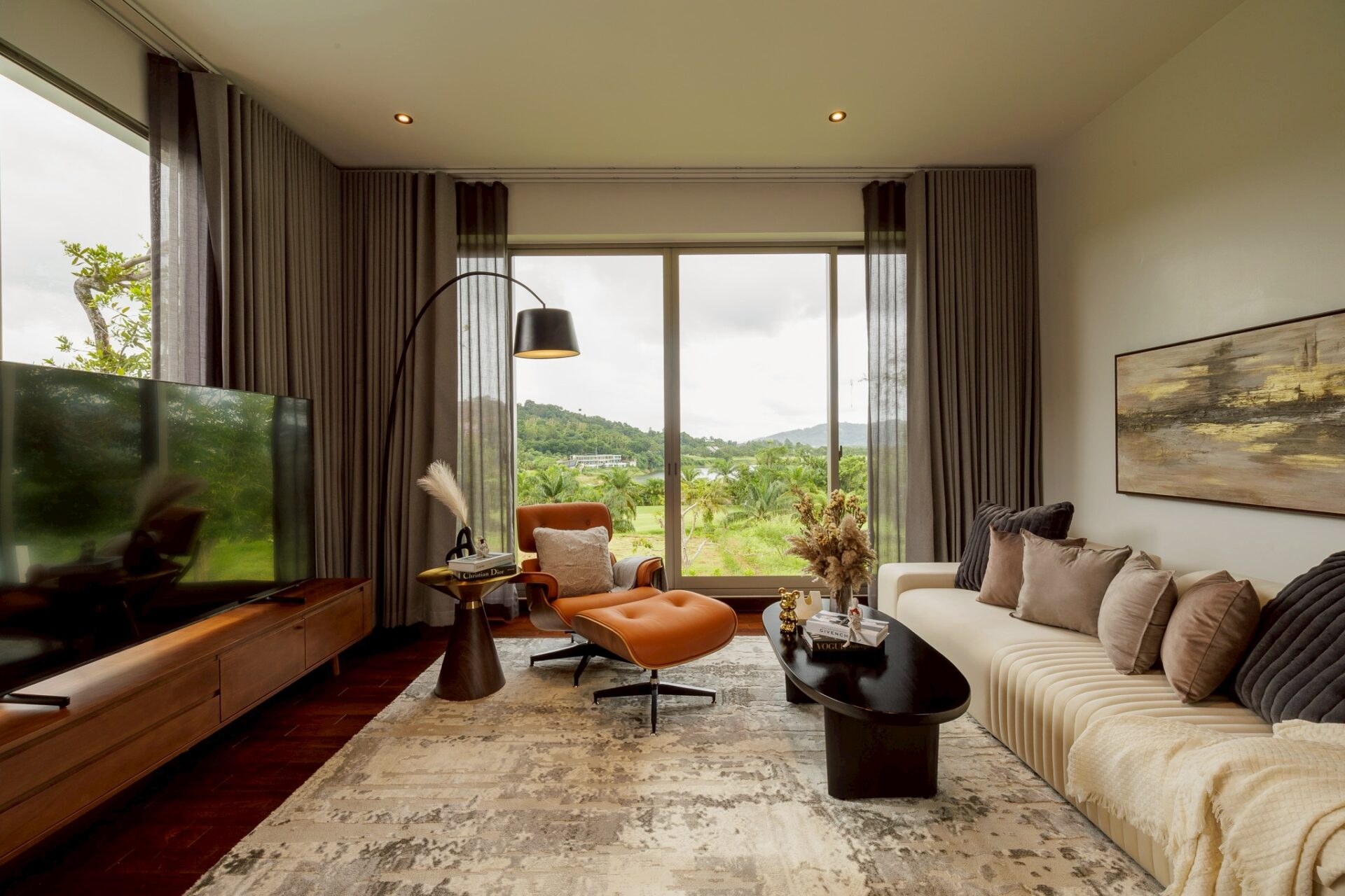 Modern living room with a beige sectional sofa, gray cushions, and large windows showing green hills outside on a sunny day.