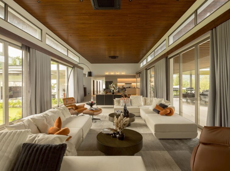 Open-plan living room with white sectional sofas, brown leather chairs, and grey curtains along floor-to-ceiling glass walls; wood-paneled ceiling and an open kitchen in the back.
