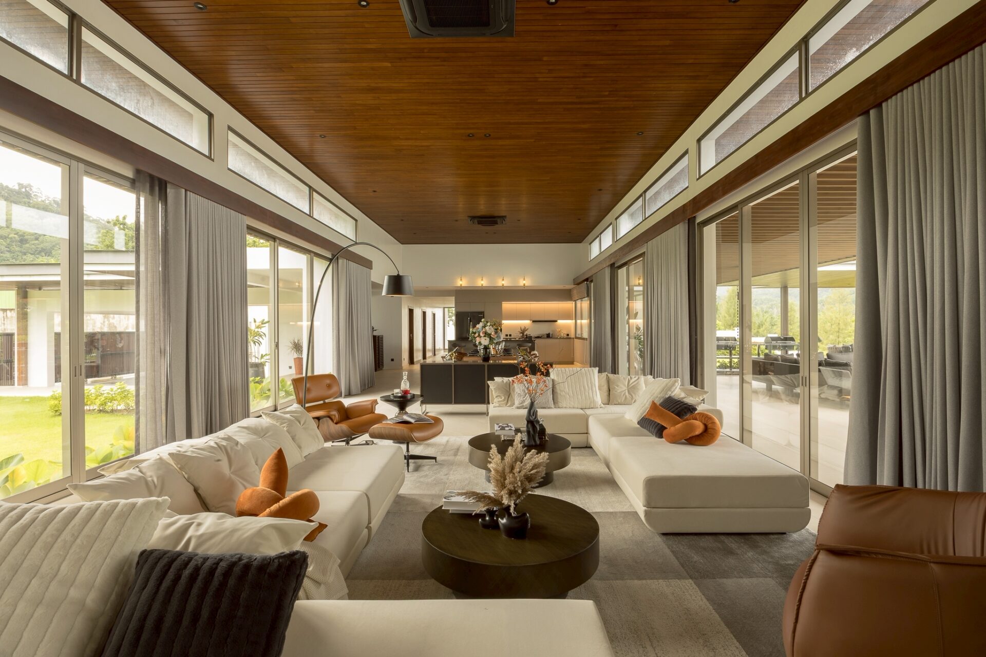 Open-plan living room with white sectional sofas, brown leather chairs, and grey curtains along floor-to-ceiling glass walls; wood-paneled ceiling and an open kitchen in the back.