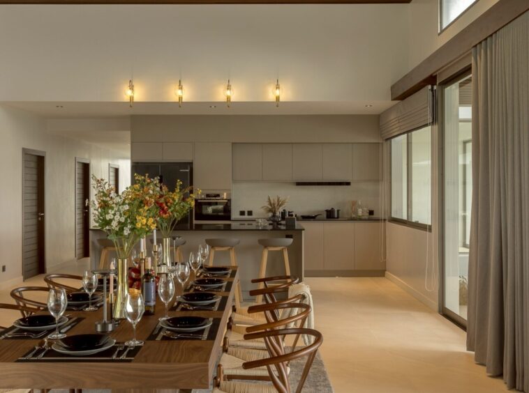 Open dining area with a long wooden table set for a formal dinner, flowers, and wine, with a modern kitchen in the background.