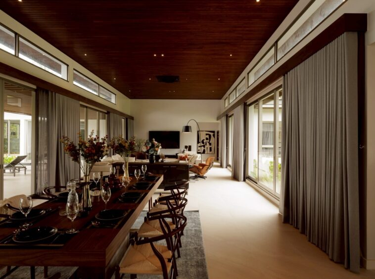Open-concept dining room with a long dark-wood table set for a formal meal, glass walls, and floor-to-ceiling curtains.