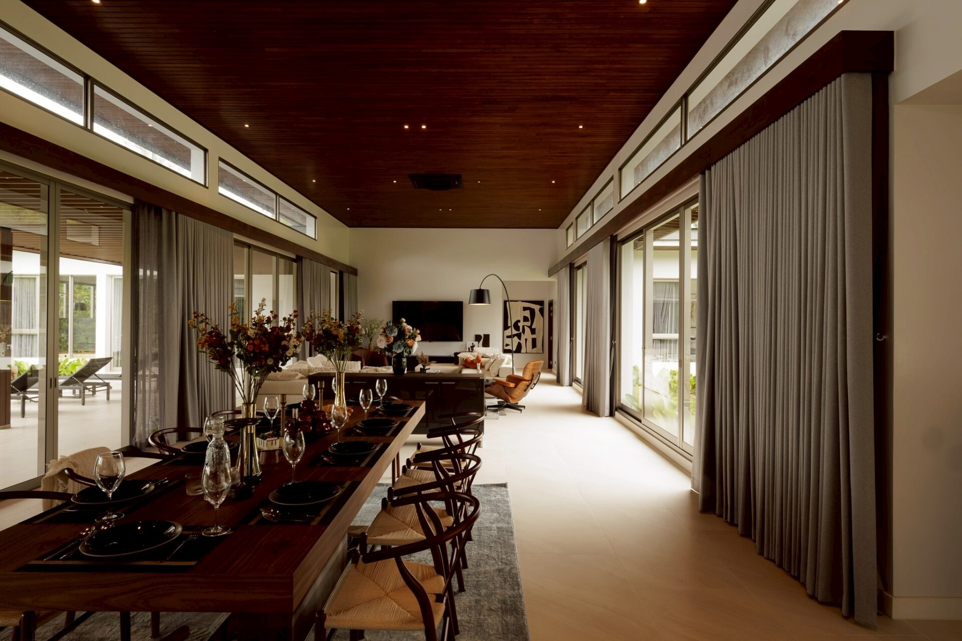 Open-concept dining room with a long dark-wood table set for a formal meal, glass walls, and floor-to-ceiling curtains.