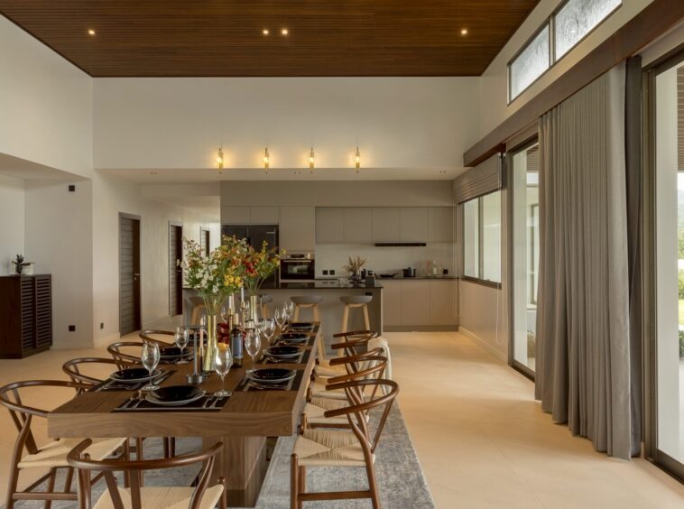Open-plan dining room with a long wooden table set for a formal meal; in the background a modern kitchen and large windows with heavy gray curtains to the right.