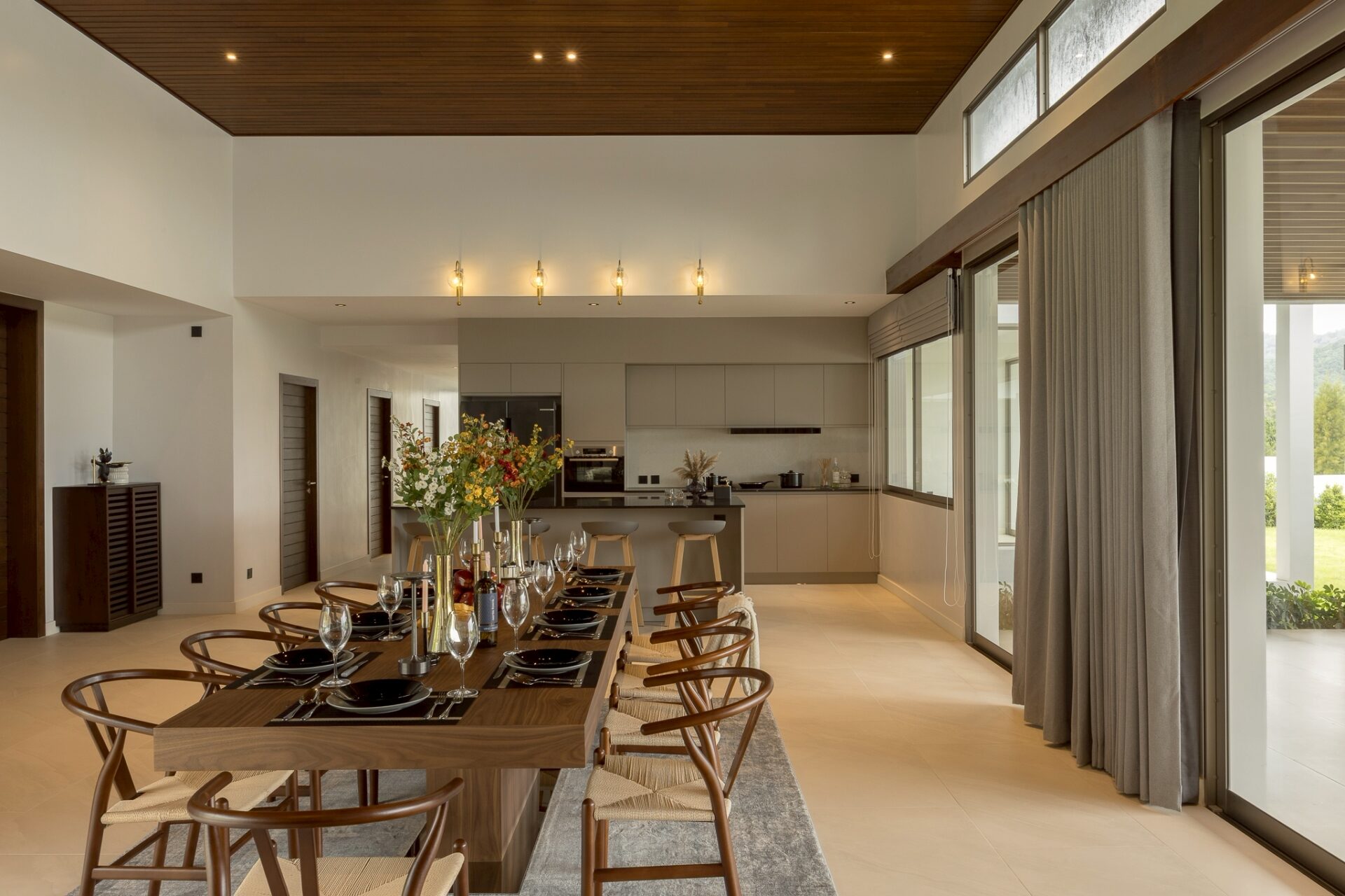 Open-plan dining room with a long wooden table set for a formal meal; in the background a modern kitchen and large windows with heavy gray curtains to the right.