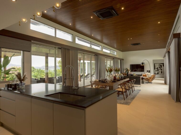 Open-plan kitchen and dining room with a dark island, stainless sink, wooden ceiling, and large windows overlooking greenery outside.