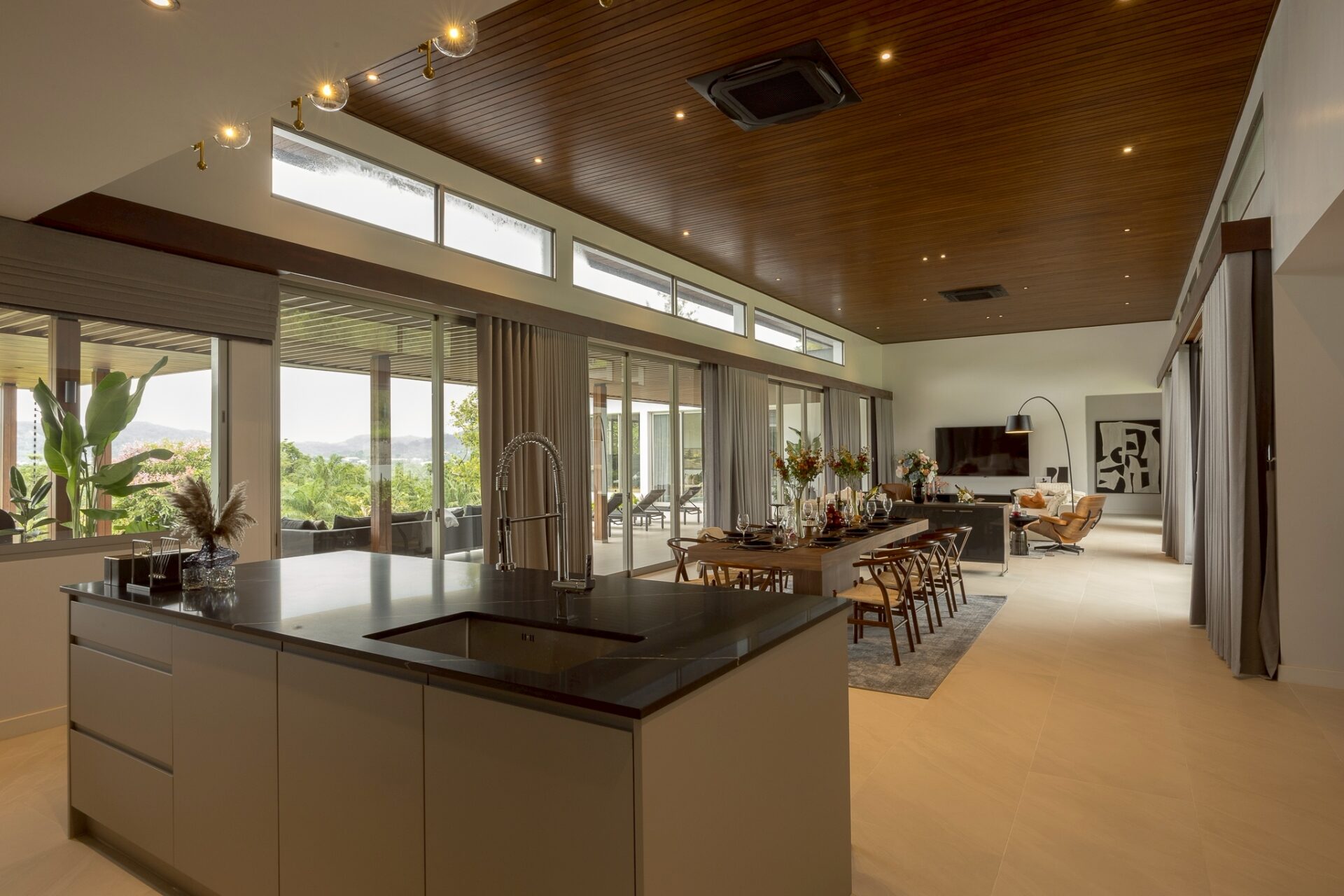 Open-plan kitchen and dining room with a dark island, stainless sink, wooden ceiling, and large windows overlooking greenery outside.