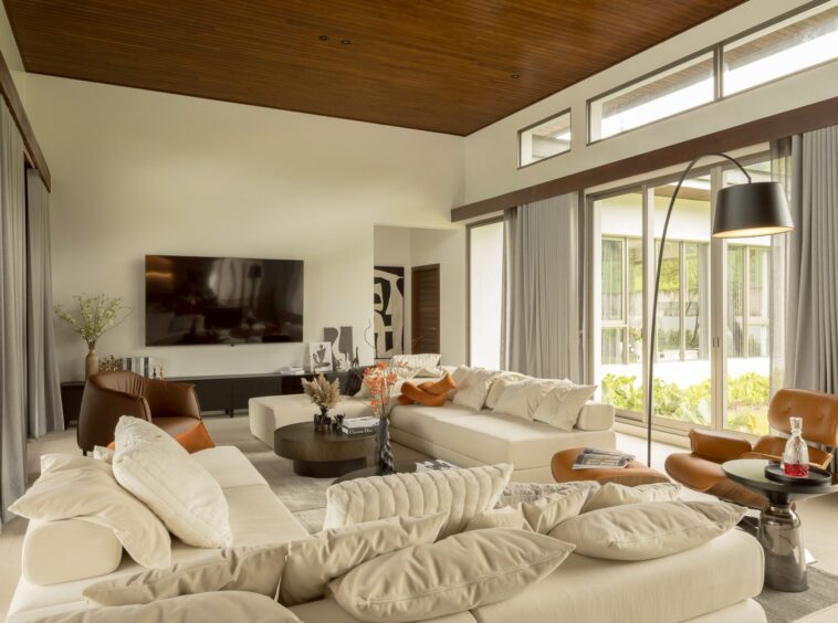 Bright, modern living room with white sectional sofa, orange cushions, and glass coffee tables facing a wall-mounted TV in a sunlit space with large windows and curtains.