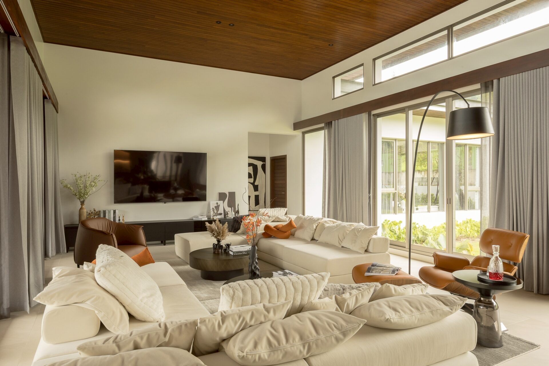 Bright, modern living room with white sectional sofa, orange cushions, and glass coffee tables facing a wall-mounted TV in a sunlit space with large windows and curtains.