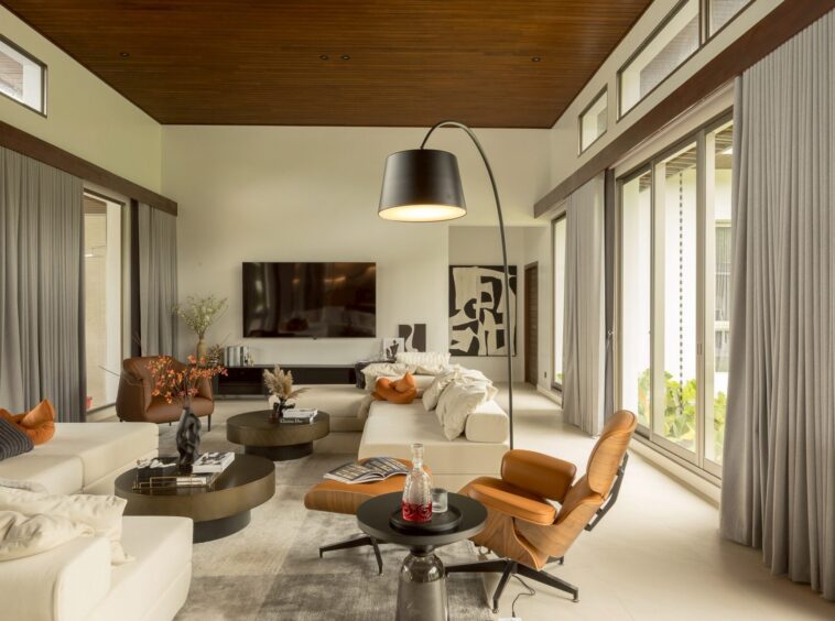 Bright modern living room with white sofas, orange pillows, and round black coffee tables under a wooden ceiling; large windows with gray curtains provide natural light.