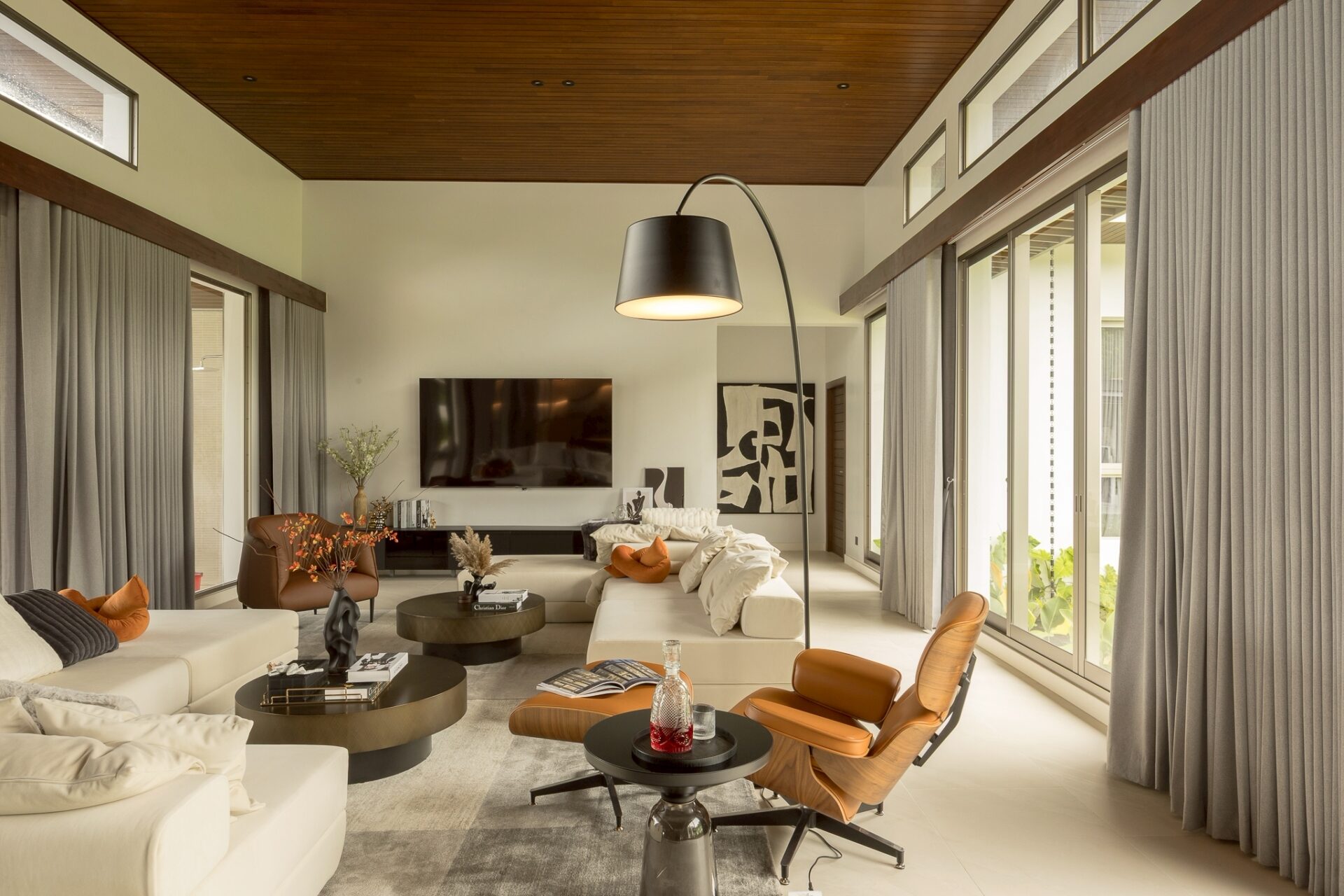 Bright modern living room with white sofas, orange pillows, and round black coffee tables under a wooden ceiling; large windows with gray curtains provide natural light.