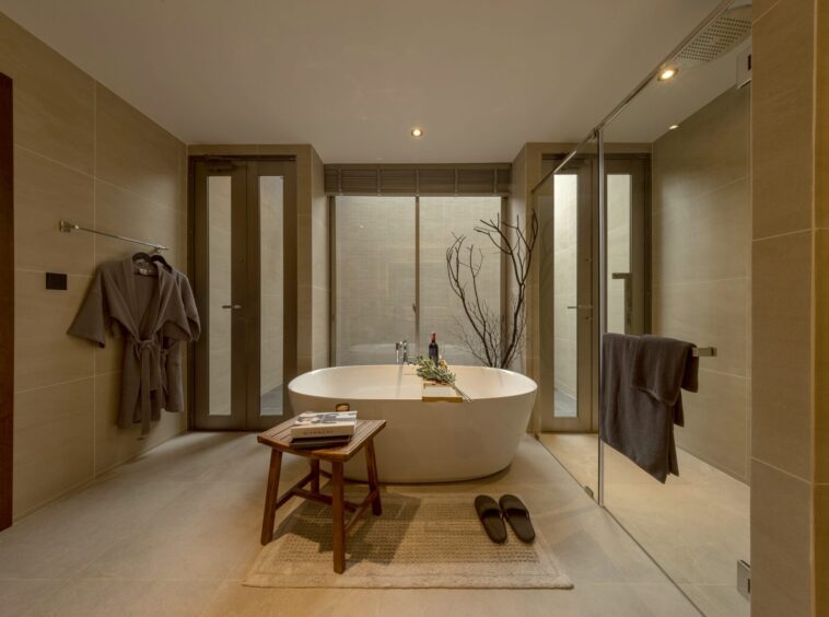 Modern spa bathroom with a freestanding tub on a rug, a wooden stool with magazines, and a glass-walled shower.