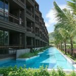 Long rectangular pool beside a dark brown building with balconies and palm trees along the edge.