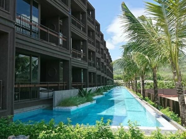 Long rectangular pool beside a dark brown building with balconies and palm trees along the edge.