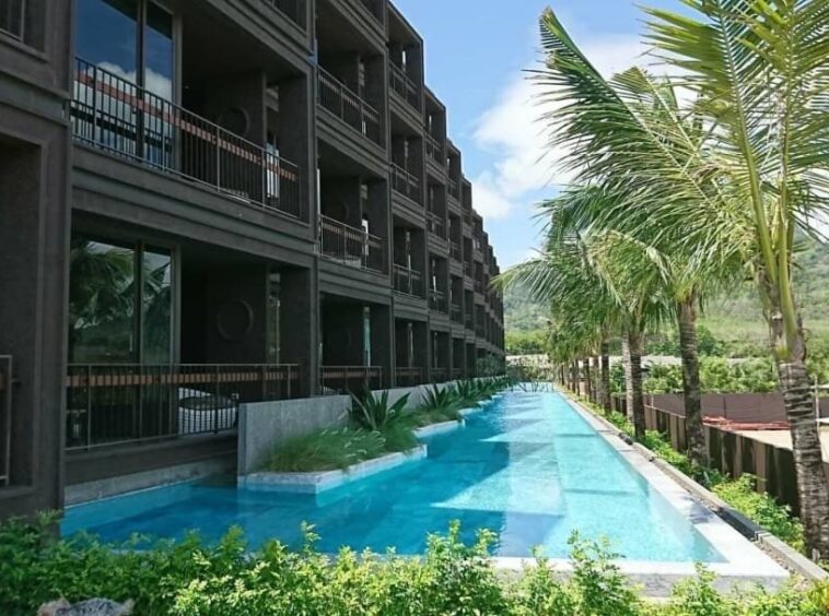 Long rectangular pool beside a dark brown building with balconies and palm trees along the edge.