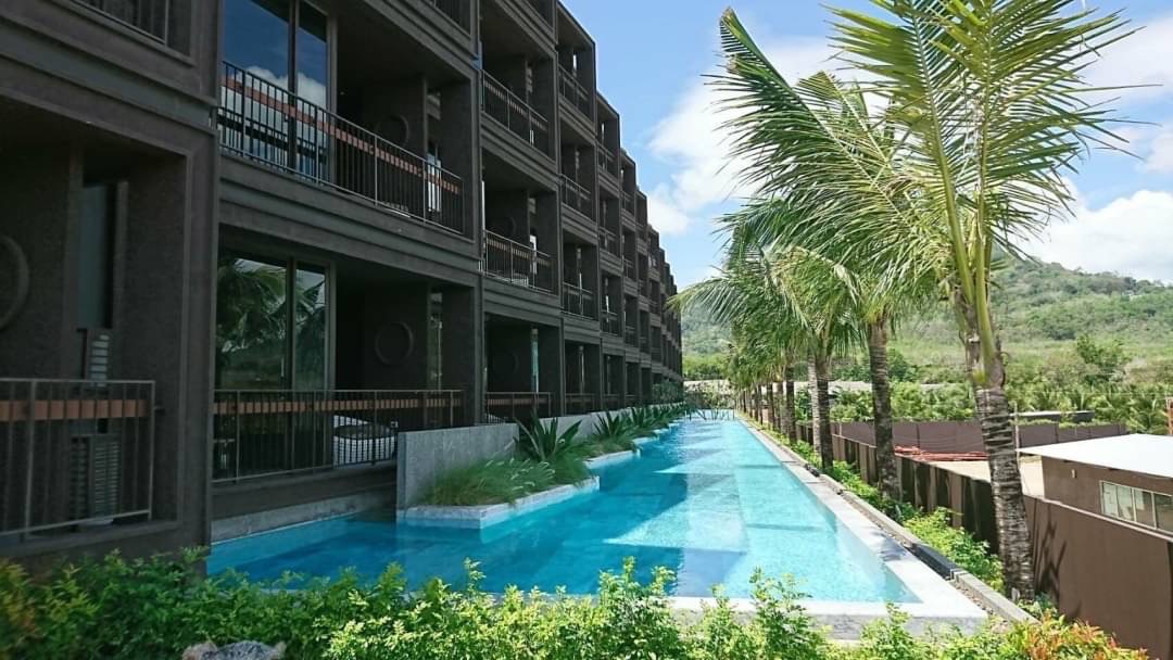 Long rectangular pool beside a dark brown building with balconies and palm trees along the edge.