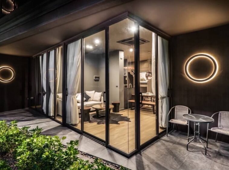 Contemporary glass-walled living room at night, featuring a sectional sofa, wooden coffee tables, and a visible kitchen, accented by circular wall lights outside.