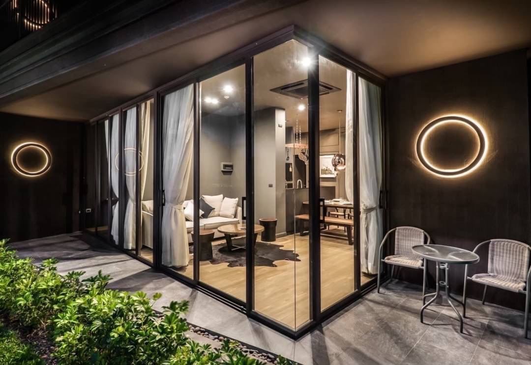 Contemporary glass-walled living room at night, featuring a sectional sofa, wooden coffee tables, and a visible kitchen, accented by circular wall lights outside.