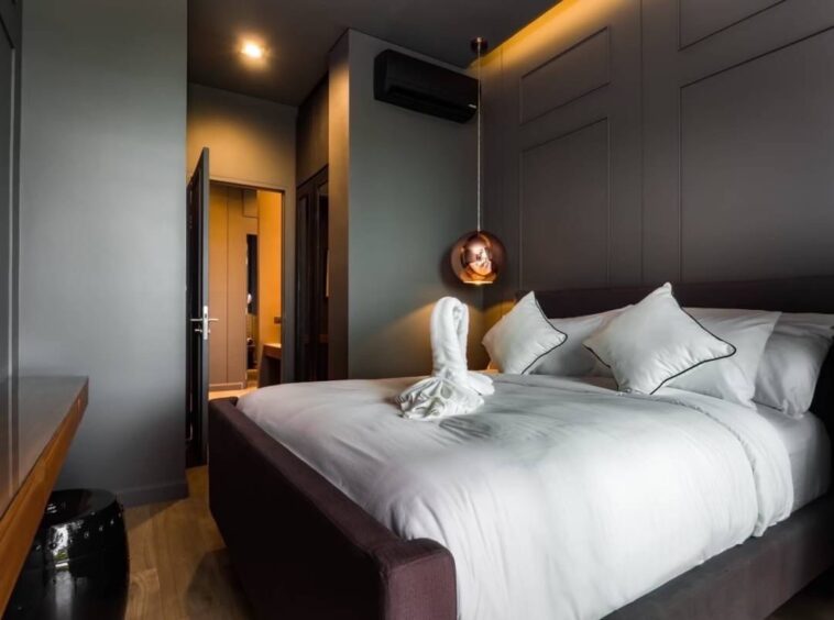 Modern hotel bedroom with a large white bed, gray walls, and a copper pendant light over the side; towel folded into a swan on the blanket.