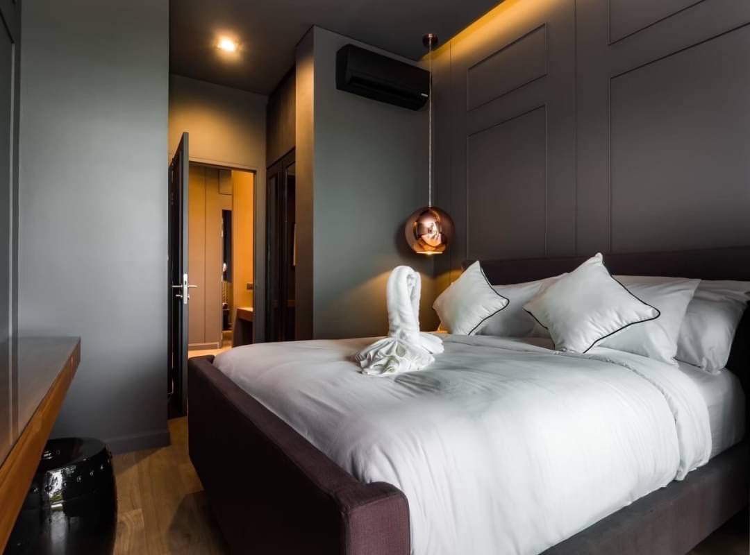 Modern hotel bedroom with a large white bed, gray walls, and a copper pendant light over the side; towel folded into a swan on the blanket.