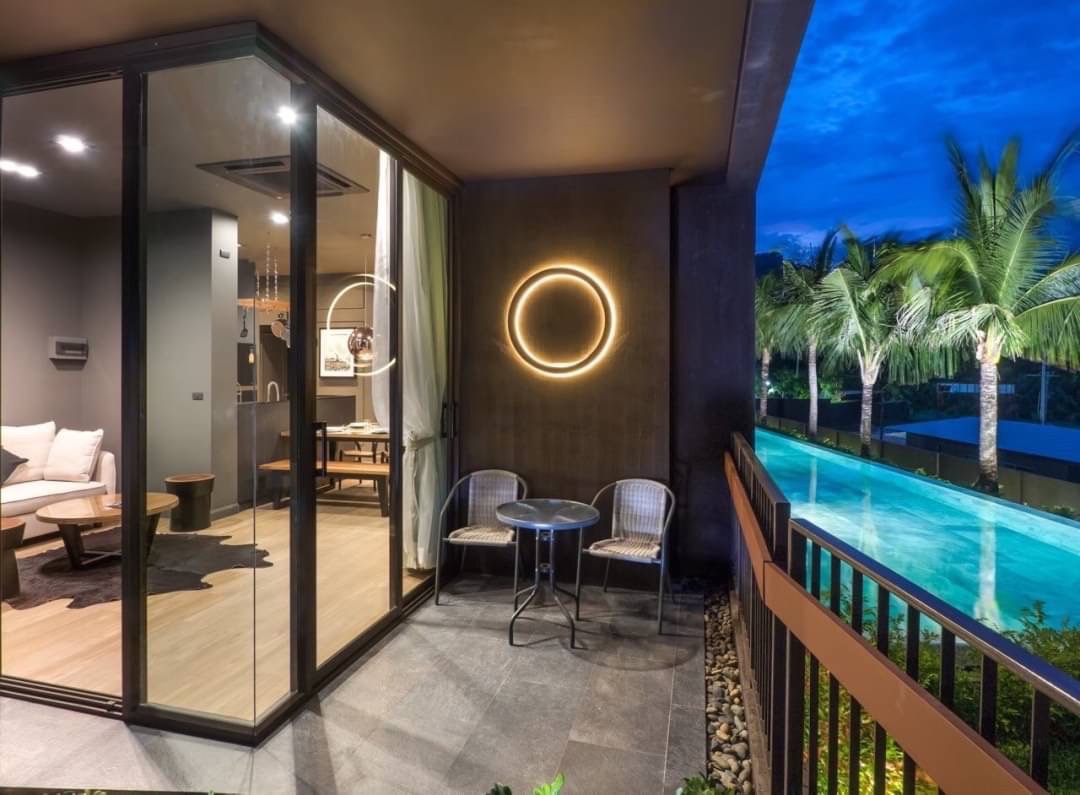 Hotel balcony at dusk with two chairs and a small table, circular wall light, and a pool with palm trees.