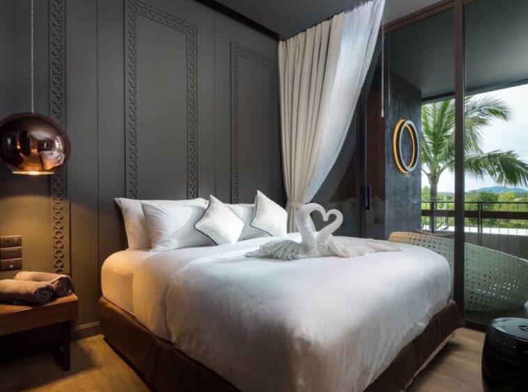 Luxurious hotel room with a large white bed, decorative gray paneled wall, and a balcony view with palm trees outside the glass door; bed has towel swans on top.