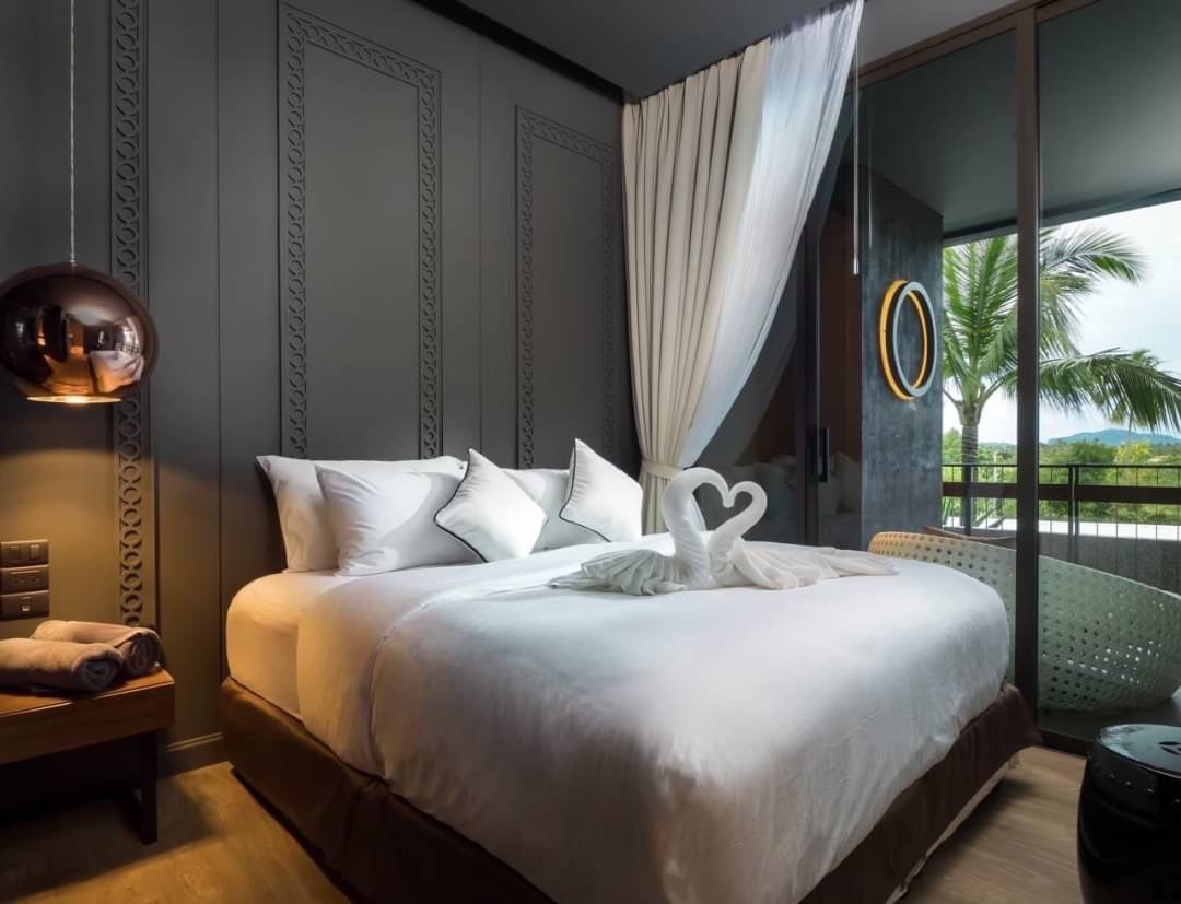Luxurious hotel room with a large white bed, decorative gray paneled wall, and a balcony view with palm trees outside the glass door; bed has towel swans on top.