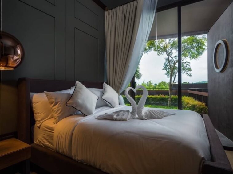 Bed with white linens and two towel swans on top, in a modern hotel room with floor-to-ceiling windows and a garden view.