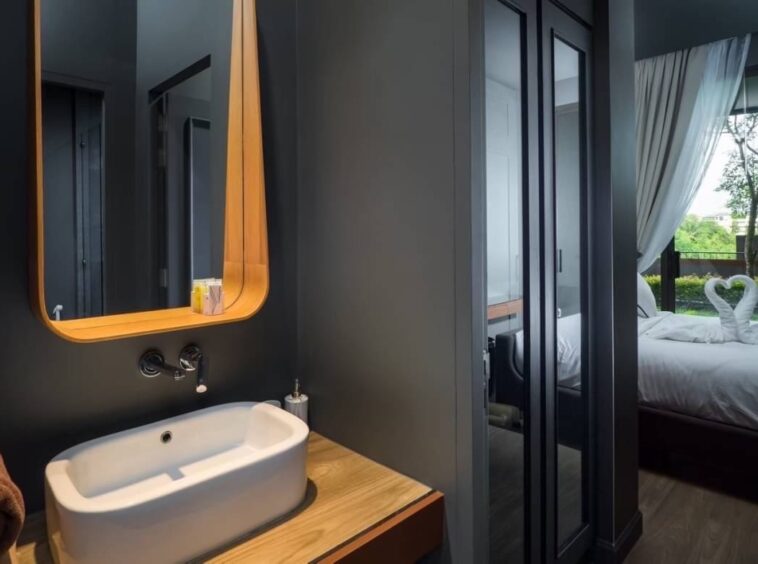 Modern bathroom: white vessel sink on a wooden counter, curved wooden mirror, gray walls, and doorway to a bedroom with swan-towel decor.