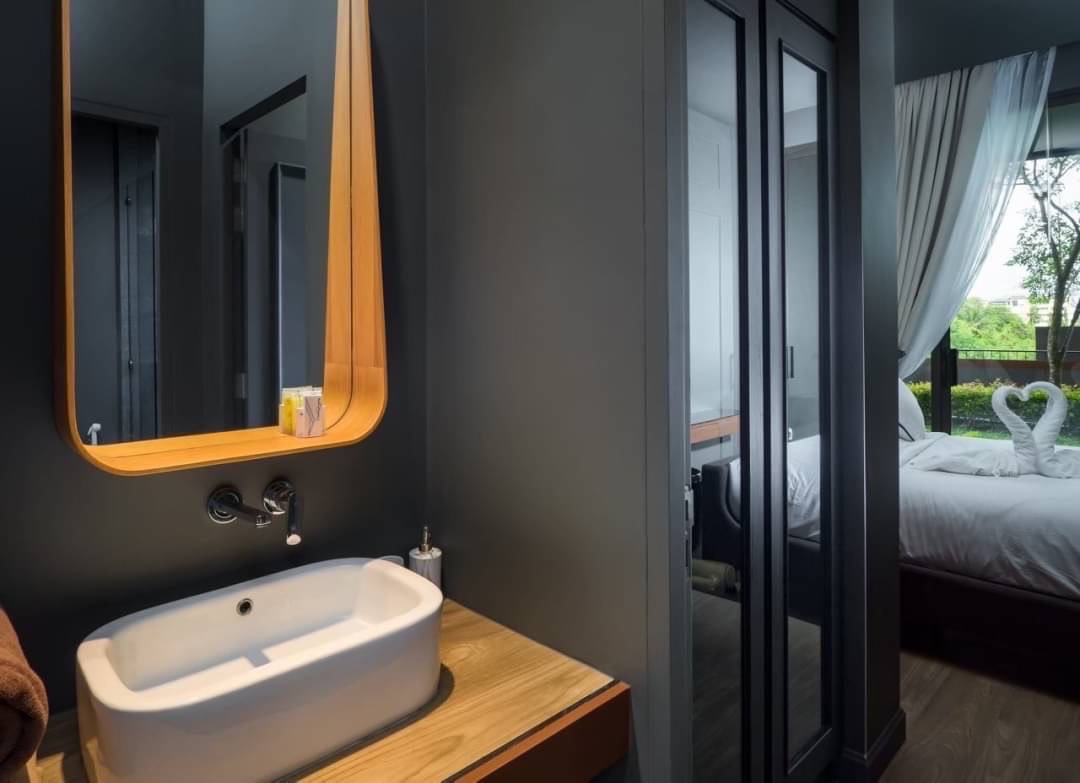 Modern bathroom: white vessel sink on a wooden counter, curved wooden mirror, gray walls, and doorway to a bedroom with swan-towel decor.