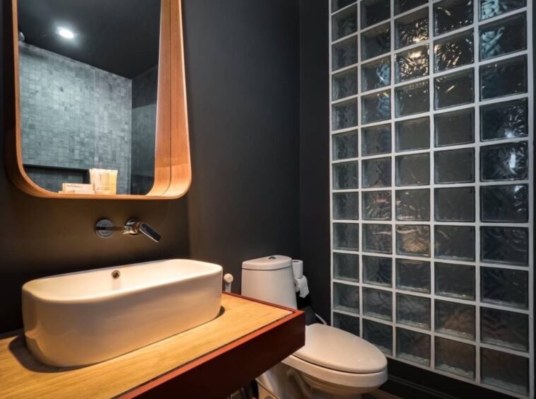 Modern bathroom with a white vessel sink on a wooden counter, round mirror, and glass block wall opposite the toilet.