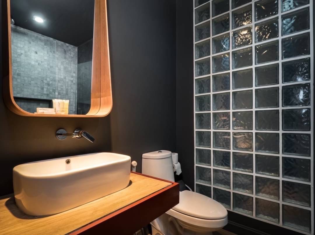 Modern bathroom with a white vessel sink on a wooden counter, round mirror, and glass block wall opposite the toilet.