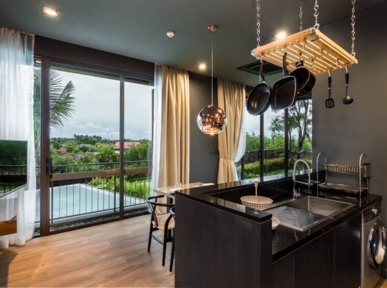 Modern kitchen with a black island, hanging pot rack, and large glass doors opening to a balcony with greenery outside.