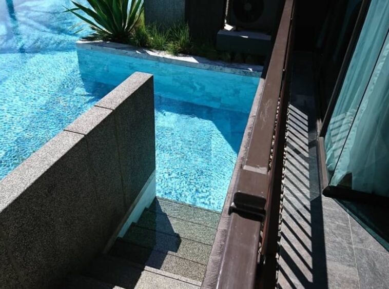 Corner of a bright blue pool with a short stone wall and steps leading into the water beside a balcony and sliding glass door.