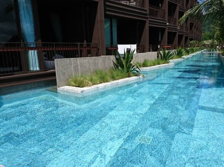 Long, luxurious outdoor hotel pool with clear blue water, palm trees, and a modern building backdrop.