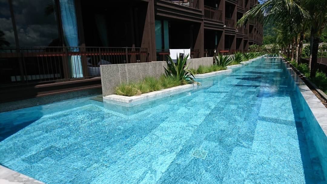 Long, luxurious outdoor hotel pool with clear blue water, palm trees, and a modern building backdrop.