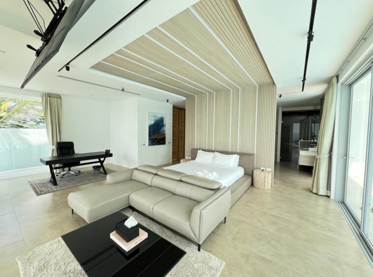 Modern hotel bedroom with a king bed against a light wood paneled accent wall, beige sofa, and a black coffee table in front of a rug.
