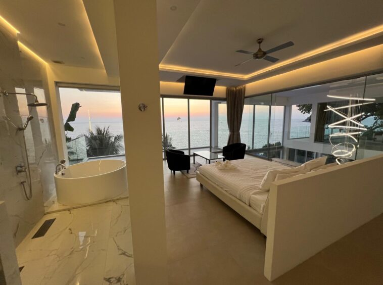 Suite with a king bed overlooking the ocean at sunset, glass walls, and a freestanding bathtub nearby.