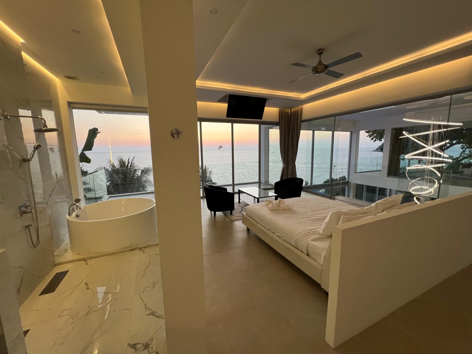Suite with a king bed overlooking the ocean at sunset, glass walls, and a freestanding bathtub nearby.