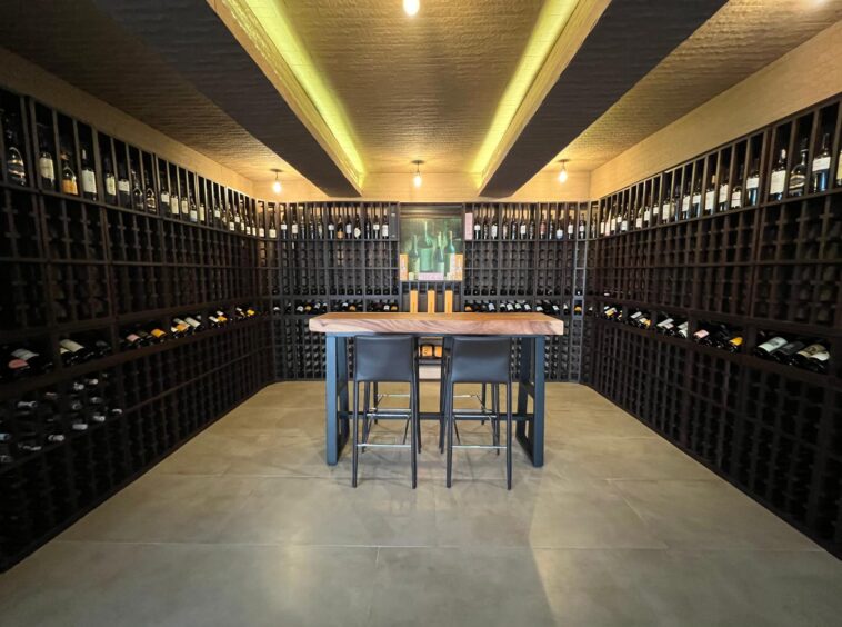 Wine cellar with dark wooden racks full of bottles surrounding a central wooden tasting table and two black stools.