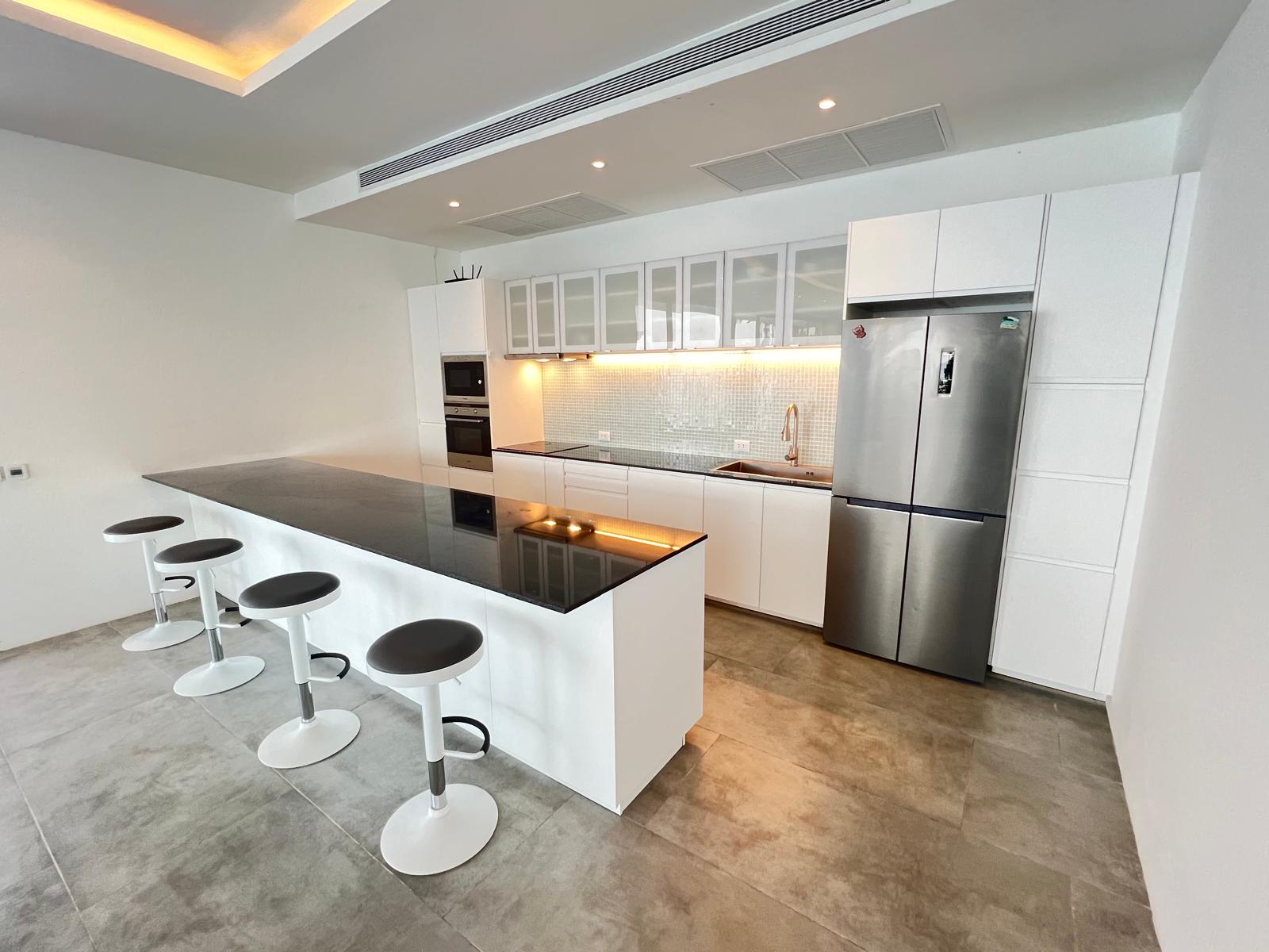 Modern white kitchen with a long black countertop island and white bar stools; stainless fridge and built-in appliances in the background