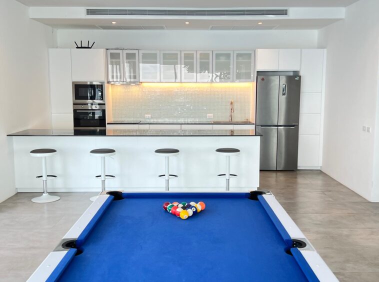 Open-plan kitchen with white cabinets, glass-front upper cabinets, and stainless steel fridge and wall ovens; a blue pool table in the foreground.
