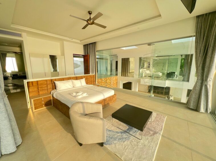 Luxurious hotel bedroom with a king bed, tan upholstered headboard, and matching nightstands next to a glass-walled living area.