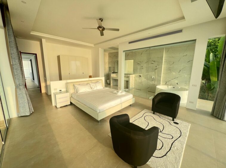 Luxurious hotel suite with white bed, black chairs, and an open marble bathroom separated by glass walls. Large windows show greenery outside.