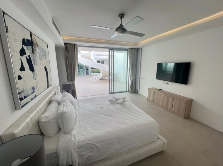 Bright hotel bedroom with a large white bed, abstract wall art, and sliding glass doors to an outdoor patio. Outside, a white spiral staircase is visible.
