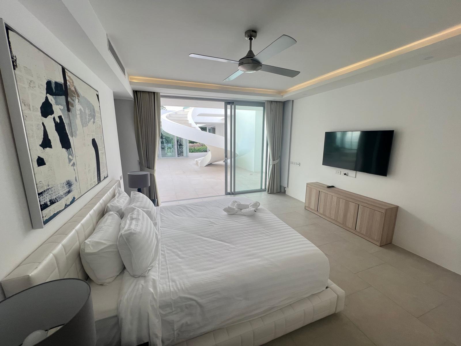 Bright hotel bedroom with a large white bed, abstract wall art, and sliding glass doors to an outdoor patio. Outside, a white spiral staircase is visible.